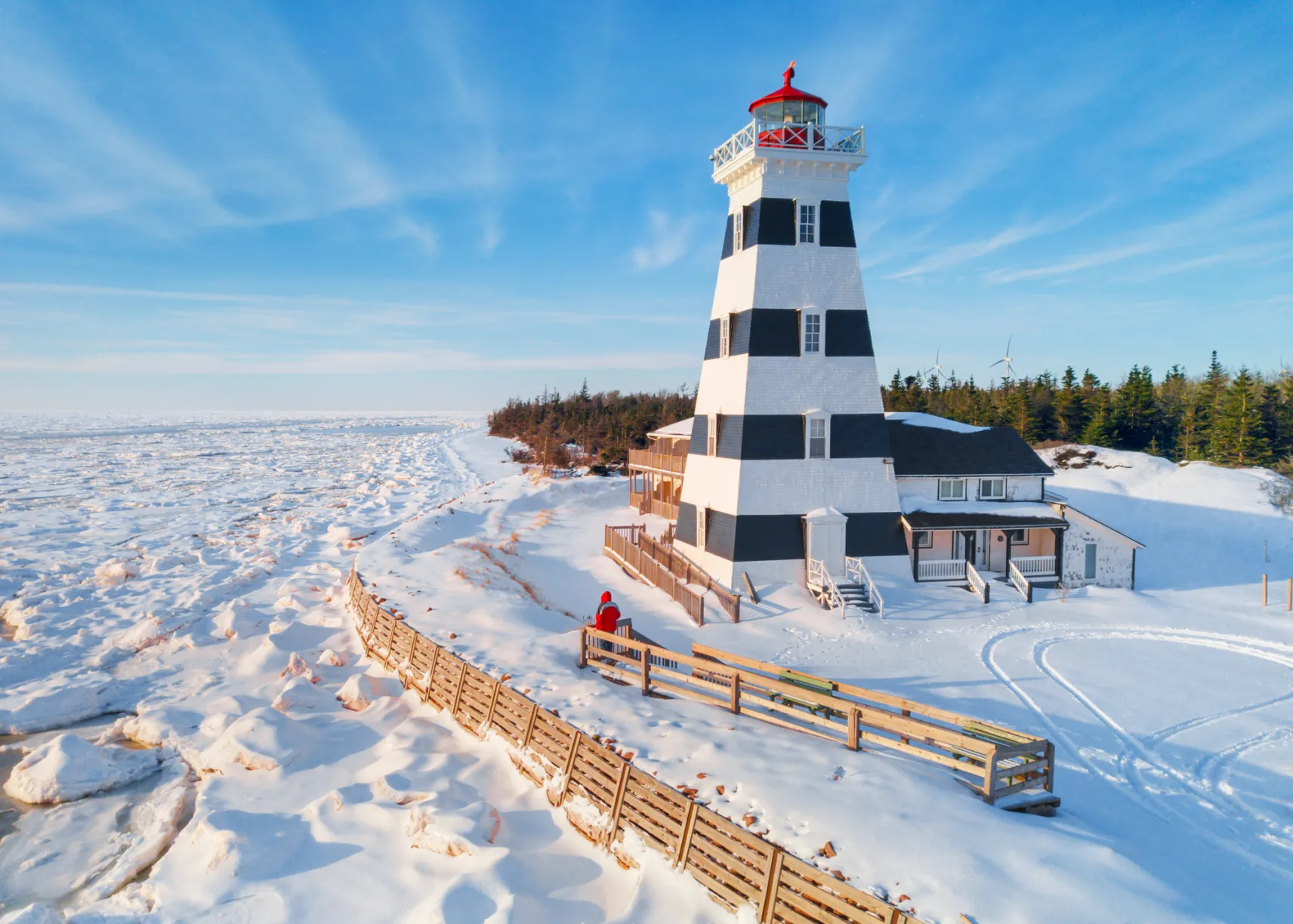 A striking black and white striped lighthouse with a red top, surrounded by deep snow and ice along a frozen shoreline under a bright blue sky.
