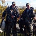Two men with goose decoys in background. One is holding a dead goose.