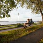 A man and a woman sit on a bench overlooking the harbour in Victoria Park in a fall setting.