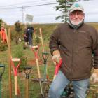 Bill Hogg stands beside a row of shovels with students planting trees in the background