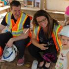 Minister Jordan Brown sits on the edge of a sandbox holding a hardhat, beside him sits Paige MacLaren, Director of the Morell Early Childhood Development Centre , and a cute little girl stands nearby.