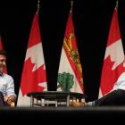 Premier Dennis King and Prime Minister Justin Trudeau sit in front of a row of Canadian flags chatting with each other.