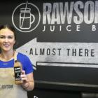 Suzanne Keough stands in front of the sign for her business, Rawsome Juice, holding jars of juices she's made.