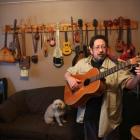 Scott Parsons is shown holding his guitar, in his home studio with guitars hanging on the wall