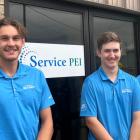 Three young men, Aaron Walsh, Johnny MacKenzie, Brandon MacQuillan, wearing blue Net Zero t-shirts stand in front of a Service PEI sign on a brick building. 