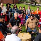 Mi'kmaq drummers, accompanied by Premier King and Mayor Brown sit drumming in a circle outside on a sunny fall day, while dancers look on. 