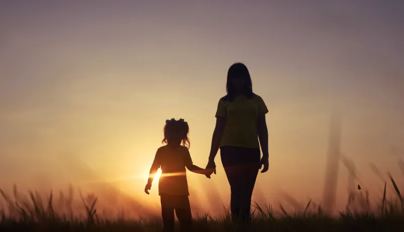 Silhouette of an adult and a child holding hands while walking through a grassy field at sunset, with the sun shining brightly behind them.