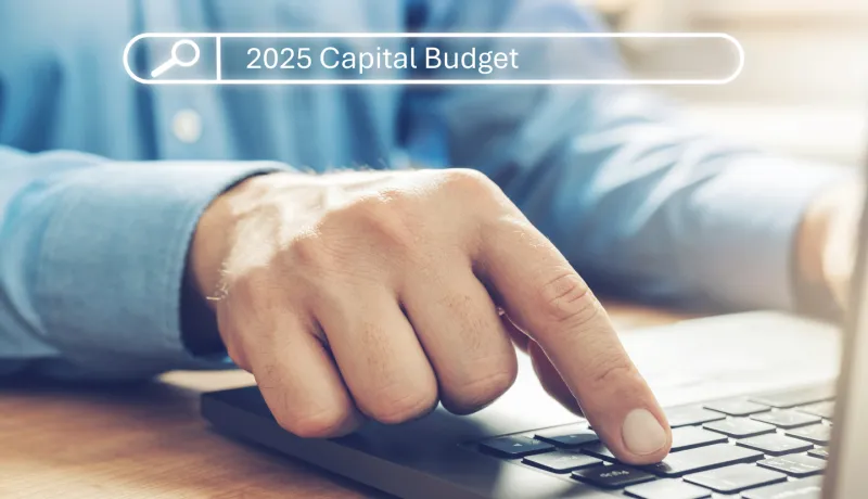 A person&#039;s hand resting on a laptop keyboard, with the screen displaying a search bar reading &quot;2025 Capital Budget&quot;