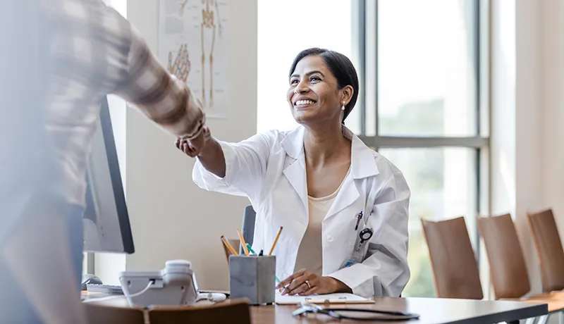 Smiling female doctor sitting at their desk greets patient by shaking their hand