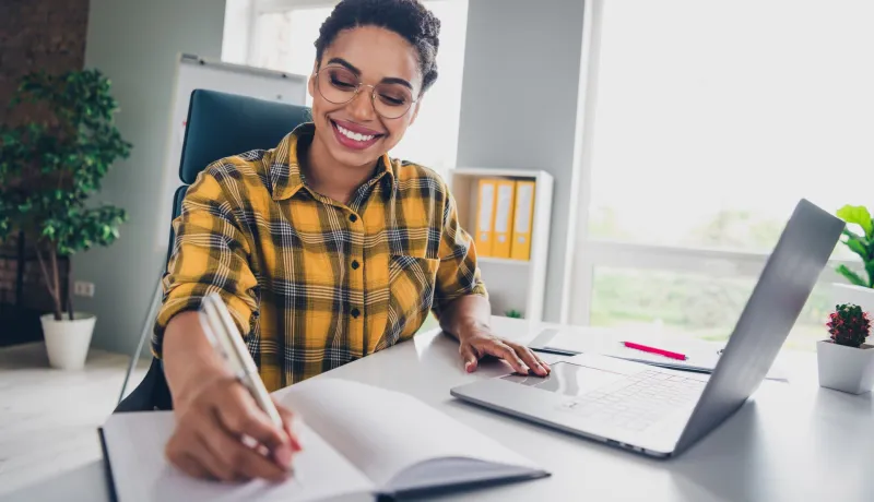 A person sitting at a desk holding a pen to a notebook and their laptop is open.