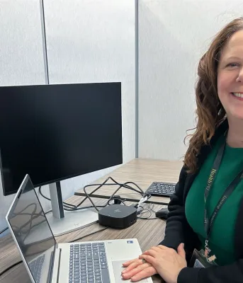 Une femme souriante aux cheveux bruns mi-longs est assise à un bureau.