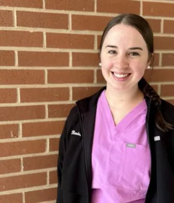A brunette young woman wearing pink scrubs smiles with a brick wall behind her.