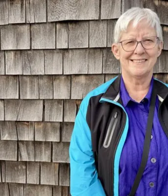A white woman with short grey hair smiles, with weathered shingles behind her.