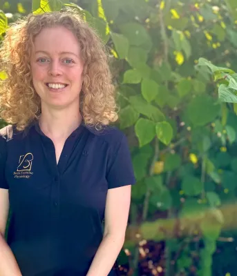 A blonde, curly-haired woman smiles in front of lush green foliage.