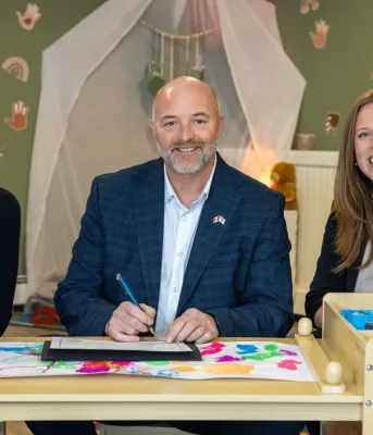 Two women and a man sit at a child's table in a playroom.