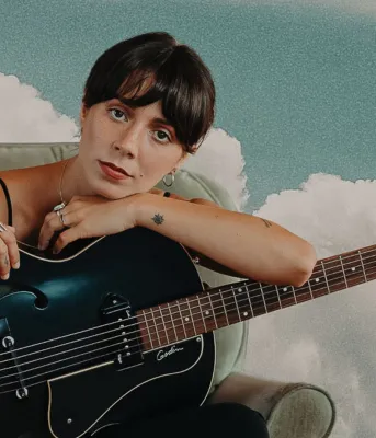 A woman holds a black guitar with blue sky and clouds behind her.