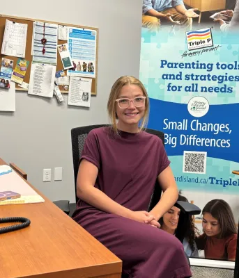 Une femme blonde est assise à son bureau avec un poster de Triple P Parenting derrière elle.