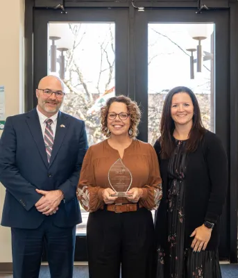 Un homme et deux femmes se tiennent côte à côte devant de grandes fenêtres.