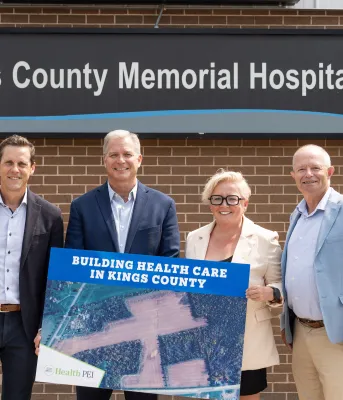 Three men and one woman stand in front of the Kings County Memorial Hospital sign while holding a poster of the newly acquired land.