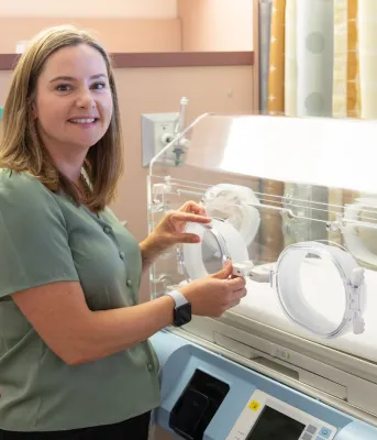 Dr. Maclean with shoulder length hair stands next to a NICU incubator.