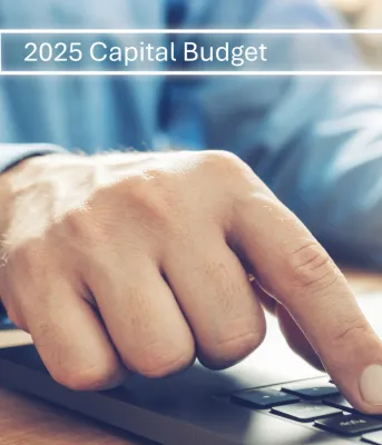 A person's hand resting on a laptop keyboard, with the screen displaying a search bar reading "2025 Capital Budget"