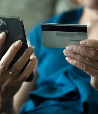 An elderly woman holds a cell phone in one hand, and a credit card in the other.