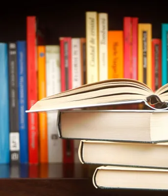 A stack of books set in front of a colourful row of books on a shelf.
