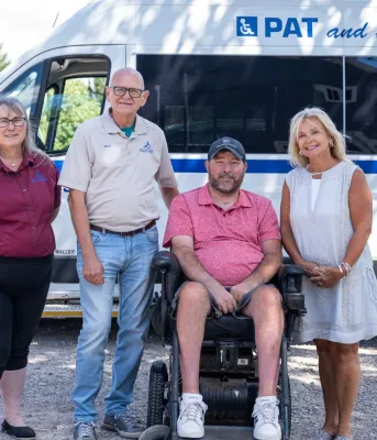 Three people stand around a man in a wheelchair in front of a Pat and the Elephant van.