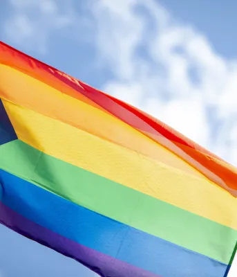 The pride flag blows in the wind with a bright blue sky in the background.