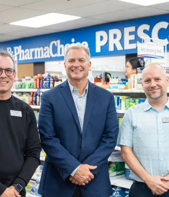Two male pharmacists and the minister stand side by side in front of a pharmacy counter.