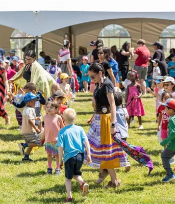 Children and adults gathered together infront of a white tent for a Pow Wow in PEI.