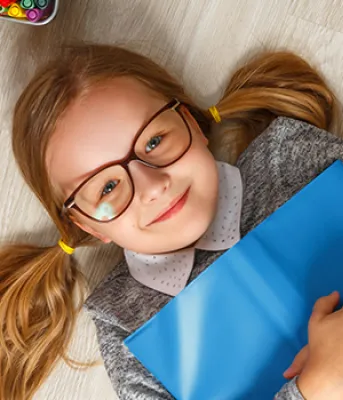Young blonde girl with glasses laying on her back on the floor holding a book surrounded by books, pencils, paint and paint brushes