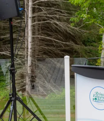 Minister Brad Trivers, sits on a stage looking at Shelley, Muzika, acting executive director of the Canadian Mental Health Association who is standing at a podium speaking.
