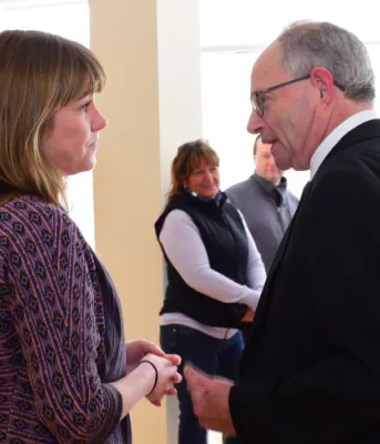 Two people facing each other talking while standing in a room filled with people.