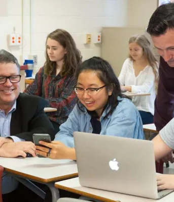 Minister Currie, a teacher and three high school students looking at a smart phone and a cumputer.