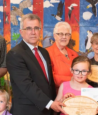 Photo shows Minister Brown surrounded by adults and children holding wooden plaques