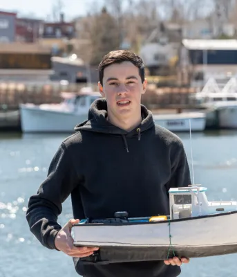 image of a person holding a model fishing boat at a wharf with fishing boats in the background