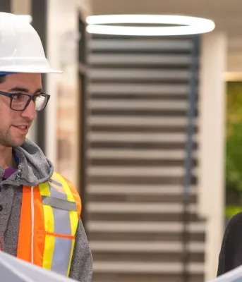 Two people, ne with a hardhat on, holding drafting documents while in a hallays.