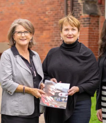 four people standing shoulder to shoulder in front of a brick building with two of them holding a booklet