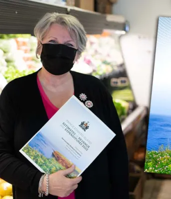 Finance Minister Darlene Compton is standing in a local grocery market holding a copy of the budget