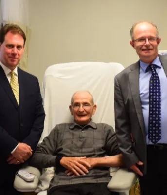 Dialysis patient Richard Fleming is in a treatment bed, with Cheryl Bank Minister James Aylward, Minister Ernie Hudson, and Irma Fleming standing nearby