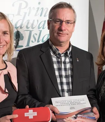 Photo shows Dr. Heather Morrisson holding a Naxolone kit, Minister Robert Henderson holding the plan, and Karen MacDonald of Justice and Public Safety.