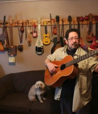 Scott Parsons is shown holding his guitar, in his home studio with guitars hanging on the wall