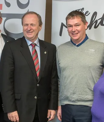 Six people standing side by side in a line in front of banners and flags