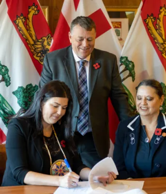 image of two people aitting at a table signing an agreement with another person standing behind them watching with some flags in the background