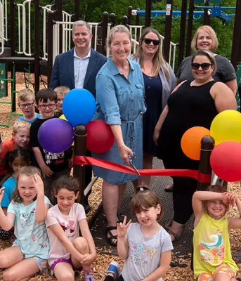 image of a group of younger and older people  cutting an ribbon in front of new playground equipment