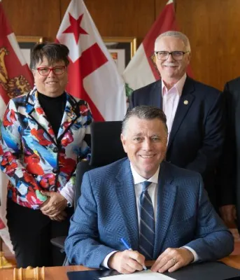 image of four people standing behind and overlooking a person while someone sits at the desk signing a proclamation