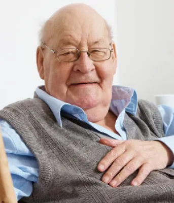 An elderly man with a cheerful expression sits comfortably in a chair, holding a cane in his right hand,
