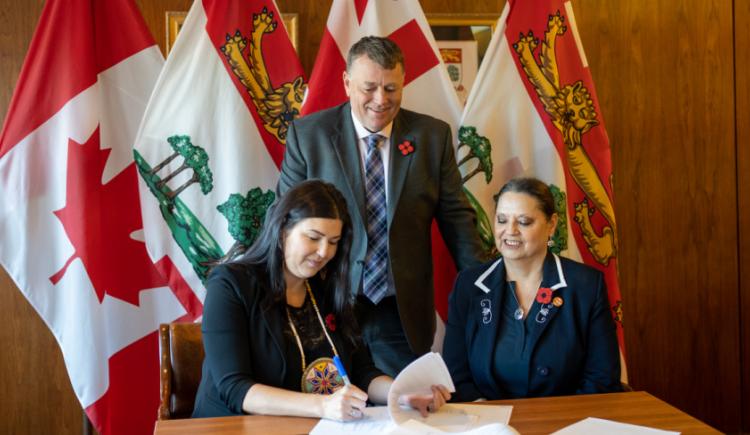 image of two people aitting at a table signing an agreement with another person standing behind them watching with some flags in the background
